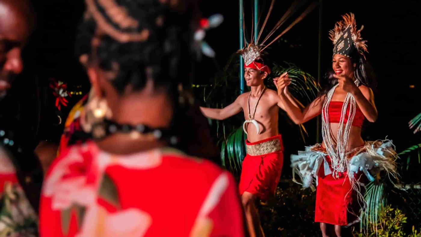 Male and female Polynesian dancers in red costumes, feathered headdresses and shell leis performing a traditional island dance at a nighttime luau