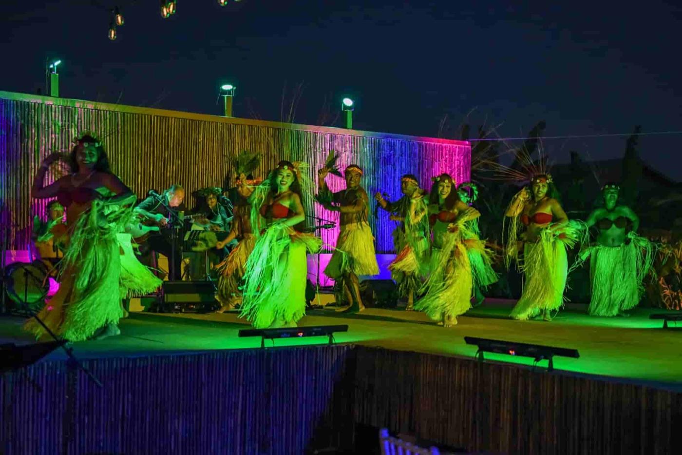 Full Polynesian revue of hula dancers in ti leaf skirts and red tops performing on a bamboo stage with live musicians under colorful lights