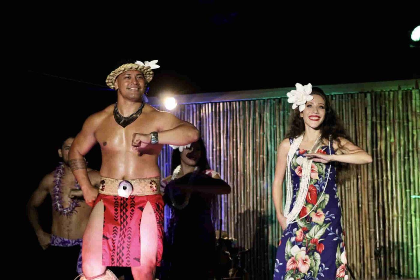 Male Polynesian dancer in red lavalava and woven headpiece performs beside a hula dancer in a floral holokū dress on a bamboo-backed luau stage at night