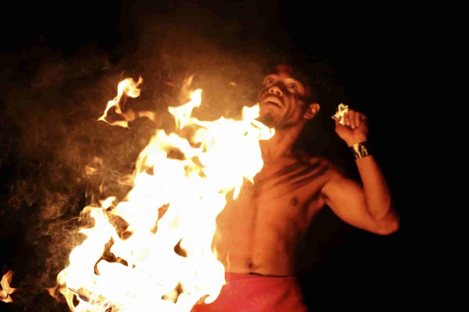 Polynesian fire knife dancer in red lavalava spinning towering flames against a black night sky during a live Hawaiian luau performance