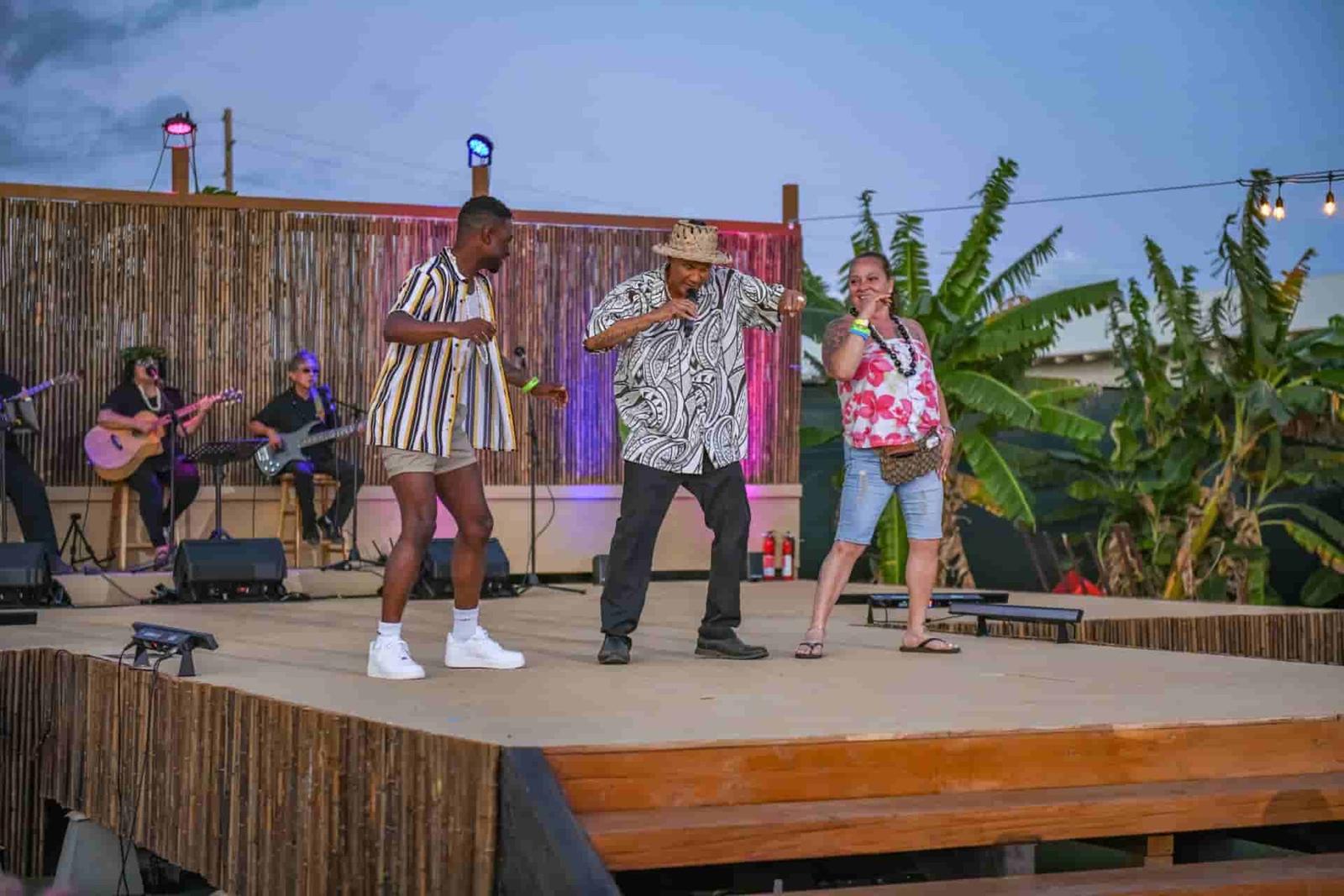 Luau emcee in aloha shirt and lauhala hat leads two smiling guests in a hula dance contest on an open-air stage as live musicians play at dusk